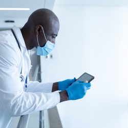 African american male doctor in hospital corridor wearing face mask using tablet. medical professional at work during coronavirus covid 19 pandemic. African american male doctor in hospital corridor wearing face mask using tablet. medical professional at work during coronavirus covid 19 pandemic.