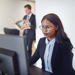 African American businesswoman working on a computer in an office Focused young African American businesswoman working on a computer at her office desk with a colleague in the background